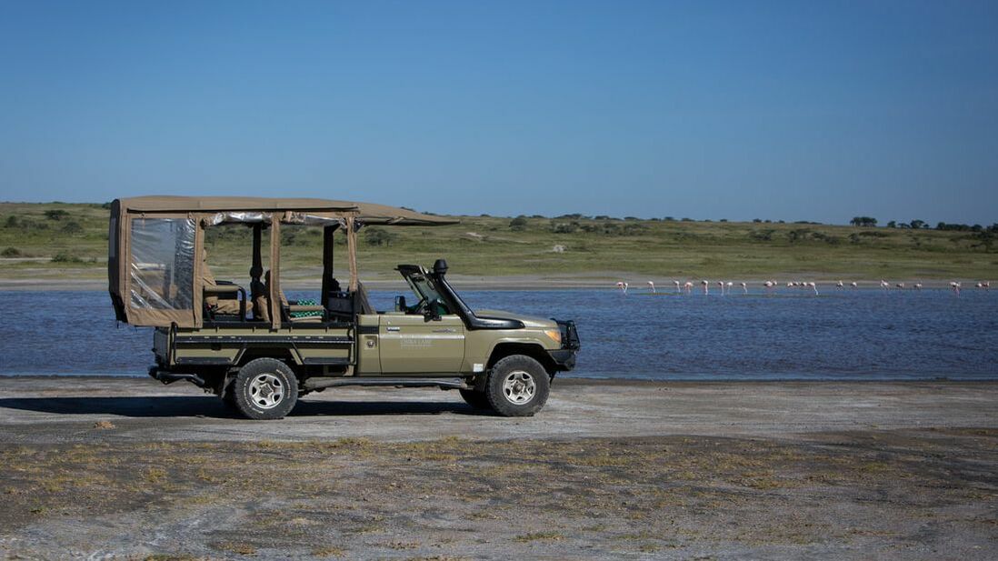 Ein Safarifahrzeug am Ufer eines Flusses in dem Flamingos sind